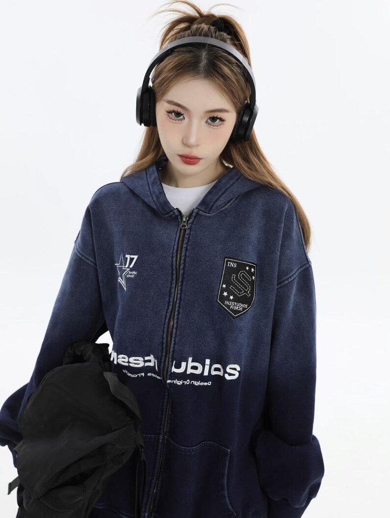 May include: A dark blue zip-up hoodie with white text and patches. The hoodie features a faded design, a front zipper, and a hood. The patches include the text "INS" and "INSTUDIOS FIRST".
