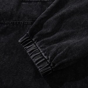 May include: Close-up of a black denim jacket sleeve. The sleeve features a ribbed cuff and a visible seam detail. The fabric has a textured appearance, with a slightly faded look.