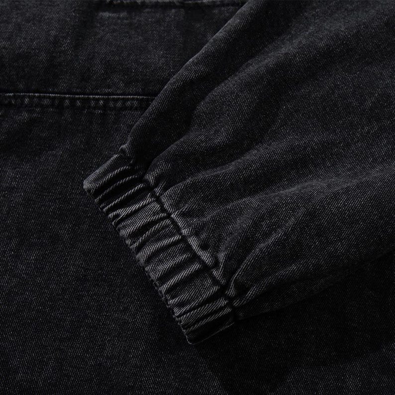 May include: Close-up of a black denim jacket sleeve. The sleeve features a ribbed cuff and a visible seam detail. The fabric has a textured appearance, with a slightly faded look.
