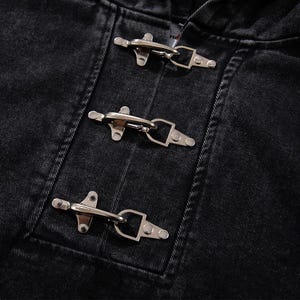 May include: Close-up of a black denim jacket with three silver toggle closures. The jacket's fabric has a textured appearance, and the metal toggles are securely fastened. The design is simple and classic.