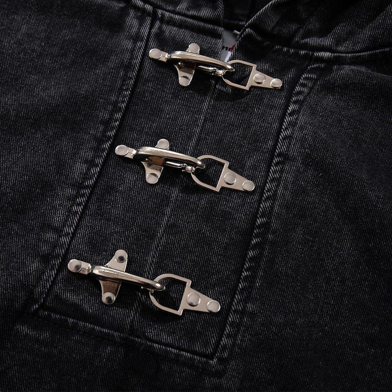 May include: Close-up of a black denim jacket with three silver toggle closures. The jacket's fabric has a textured appearance, and the metal toggles are securely fastened. The design is simple and classic.