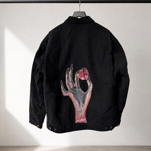 May include: Black jacket with a button-down front and a collar, hanging on a black metal rod. The back of the jacket features a graphic of a hand holding a red object. The hand is gray and red, with a white background.