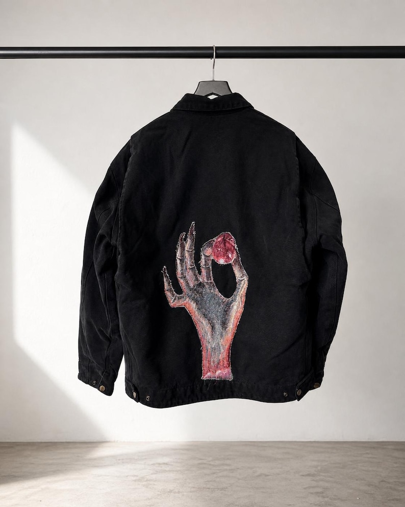 May include: Black jacket with a button-down front and a collar, hanging on a black metal rod. The back of the jacket features a graphic of a hand holding a red object. The hand is gray and red, with a white background.