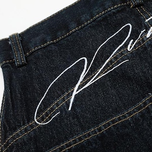 K&ouml;nnte beinhalten: Nahaufnahme einer dunkel gewaschenen Jeans mit wei&szlig; gesticktem Schriftzug auf der Ges&auml;&szlig;tasche. Die Jeans hat einen klassischen F&uuml;nf-Taschen-Stil mit kontrastierenden gelben N&auml;hten.