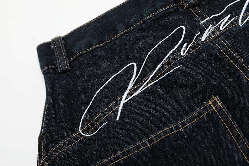 K&ouml;nnte beinhalten: Nahaufnahme einer dunkel gewaschenen Jeans mit wei&szlig; gesticktem Schriftzug auf der Ges&auml;&szlig;tasche. Die Jeans hat einen klassischen F&uuml;nf-Taschen-Stil mit kontrastierenden gelben N&auml;hten.