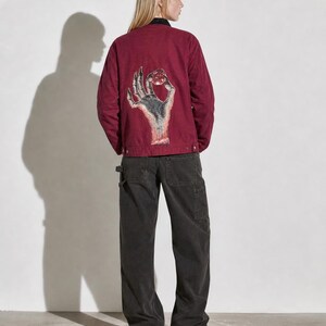 May include: Burgundy jacket with a hand graphic on the back, paired with dark gray work pants. The jacket features a black collar and button closures. The hand graphic is detailed with shades of gray and red.