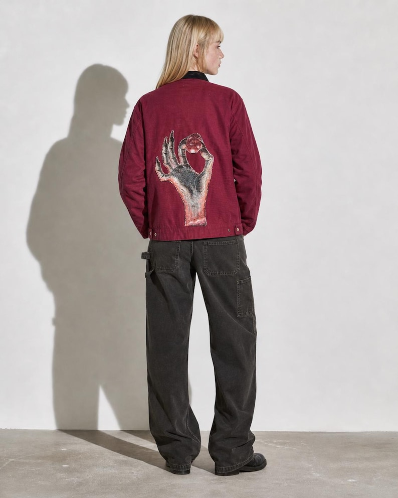May include: Burgundy jacket with a hand graphic on the back, paired with dark gray work pants. The jacket features a black collar and button closures. The hand graphic is detailed with shades of gray and red.
