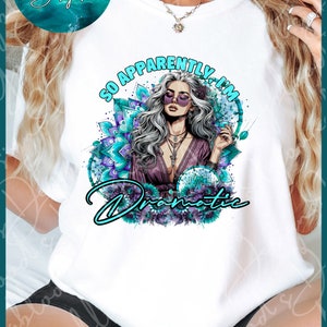 Könnte beinhalten: Weißes T-Shirt mit einem Grafikdesign einer Frau mit langen grauen Haaren, die eine Sonnenbrille und ein lila Kleid trägt. Die Grafik ist von türkisfarbenen und violetten Blumendesigns umgeben und der Text "So Apparently I'm Dramatic".
