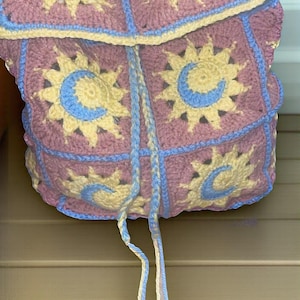 May include: A crocheted backpack with a pink, yellow, and blue color scheme. The backpack features a repeating pattern of suns and moons.