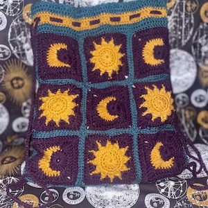 Op de afbeelding: Gehaakte tas met een hemels thema. De tas is voornamelijk paars met gele zonnen en halve manen in vierkanten. Blauwgroene en gouden afwerking. De tas heeft trekkoorden.