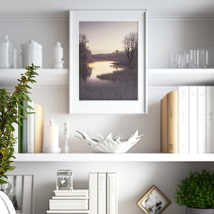 May include: White bookshelf with three shelves, featuring a framed landscape photograph, decorative items, and books. The top shelf has a glass bowl, two white ceramic jars, and a small white ceramic jar with a lid. The middle shelf has a white ceramic candle, a white ceramic figurine, a white ceramic bowl, and a stack of books. The bottom shelf has a stack of books, a small glass lantern, a framed photograph, and a small potted plant.