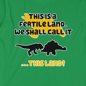 Puede incluir: Camiseta verde con un gráfico negro y amarillo de dos dinosaurios. El texto en la camiseta dice "This is a fertile land. We shall call it... This Land!"