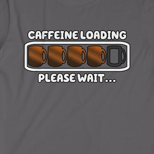 Pode incluir: Uma camiseta cinza escura com um gráfico branco de uma barra de carregamento de cafeína. A barra está cheia de cinco xícaras de café, com a última xícara vazia. O texto "CAFFEINE LOADING" e "PLEASE WAIT..." está impresso em branco.