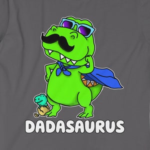 Può includere: Una maglietta grigia con un dinosauro verde che indossa occhiali da sole e un mantello blu. Il dinosauro ha i baffi e tiene un piccolo dinosauro. Il testo "DADASAURUS" è stampato sotto il dinosauro.