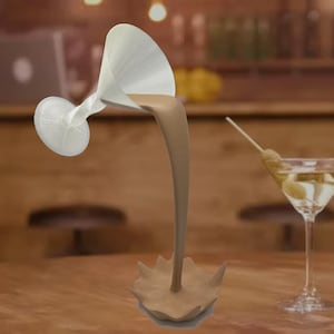 Può includere: Un bicchiere da martini bianco con un liquido marrone che cola fuori, formando uno schizzo marrone su un tavolo di legno. Un bicchiere da martini con olive è sullo sfondo.