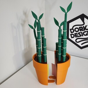 Peut inclure: Une plante de bambou d&eacute;corative imprim&eacute;e en 3D dans un pot orange. Les tiges de bambou sont vertes avec des bandes blanches, et les feuilles sont &eacute;galement vertes. Le pot est coup&eacute; en deux, r&eacute;v&eacute;lant un int&eacute;rieur jaune et rose. Le logo "Dorky Designs" est visible.