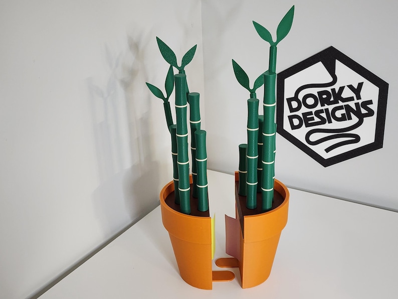 Peut inclure: Une plante de bambou d&eacute;corative imprim&eacute;e en 3D dans un pot orange. Les tiges de bambou sont vertes avec des bandes blanches, et les feuilles sont &eacute;galement vertes. Le pot est coup&eacute; en deux, r&eacute;v&eacute;lant un int&eacute;rieur jaune et rose. Le logo "Dorky Designs" est visible.