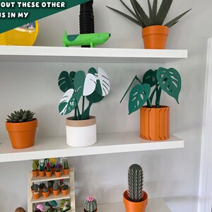 Peut inclure: Pr&eacute;sentation de sculptures de plantes en papier sur des &eacute;tag&egrave;res blanches. Les plantes sont dans des pots orange et blancs. Les plantes comprennent une succulente, un cactus et un monstera. Le texte de l'image dit "CHECK OUT THESE OTHER DESIGNS IN MY SHOP".