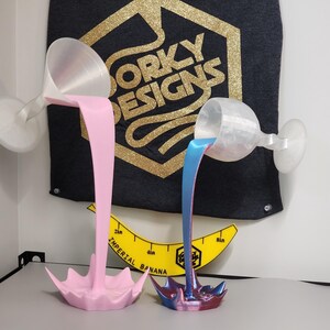Peut inclure: Deux sculptures décoratives en forme de verre à vin, une rose et une bleue, semblent verser du liquide. Les sculptures sont montées sur un mur et une table. Une bannière avec le texte "DORKY DESIGNS" est en arrière-plan.