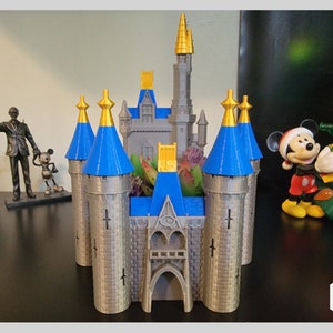 May include: A 3D printed model of Cinderella's Castle from Disney World. The castle is gray with blue and gold accents. The castle is a planter with a space for plants in the center.