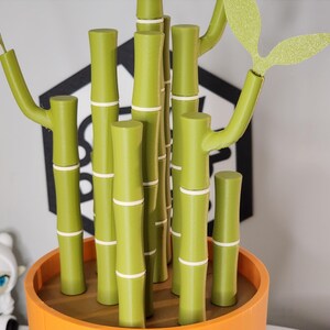 Peut inclure: Une plante de bambou d&eacute;corative dans un pot orange. Les tiges de bambou sont vertes avec des bandes blanches, et les feuilles sont vert clair avec des paillettes. L'arri&egrave;re-plan comprend une forme g&eacute;om&eacute;trique noire et blanche.