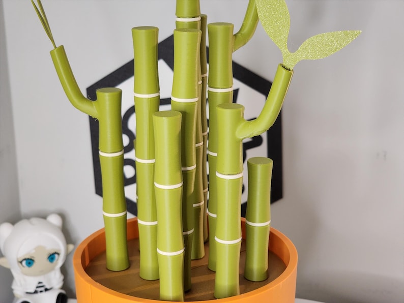 Peut inclure: Une plante de bambou d&eacute;corative dans un pot orange. Les tiges de bambou sont vertes avec des bandes blanches, et les feuilles sont vert clair avec des paillettes. L'arri&egrave;re-plan comprend une forme g&eacute;om&eacute;trique noire et blanche.