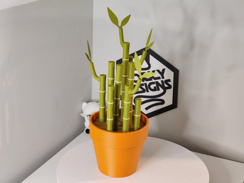 Peut inclure: Un pot orange contient une plante de bambou verte avec des d&eacute;tails blancs. Le bambou a plusieurs tiges et feuilles. Le pot est pos&eacute; sur une surface blanche. Un panneau avec le texte "SXY SIGNS" est en arri&egrave;re-plan.
