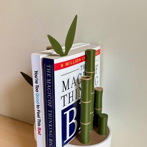 Peut inclure: Un serre-livres sur le th&egrave;me du bambou avec une base blanche. Le serre-livres pr&eacute;sente des tiges et des feuilles de bambou vertes, tenant deux livres. Les titres des livres sont "You're Too Good to Feel This Bad" et "The Magic of Thinking Big."