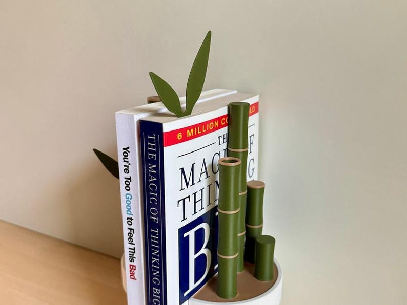 Peut inclure: Un serre-livres sur le th&egrave;me du bambou avec une base blanche. Le serre-livres pr&eacute;sente des tiges et des feuilles de bambou vertes, tenant deux livres. Les titres des livres sont "You're Too Good to Feel This Bad" et "The Magic of Thinking Big."
