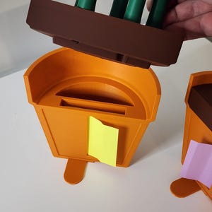 Peut inclure: Organisateur de bureau en plastique orange avec une section sup&eacute;rieure marron contenant des marqueurs verts. Un post-it jaune est attach&eacute; &agrave; l'avant. Un autre organisateur est visible &agrave; droite avec un post-it rose.