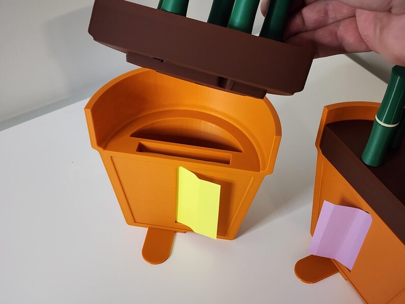 Peut inclure: Organisateur de bureau en plastique orange avec une section sup&eacute;rieure marron contenant des marqueurs verts. Un post-it jaune est attach&eacute; &agrave; l'avant. Un autre organisateur est visible &agrave; droite avec un post-it rose.
