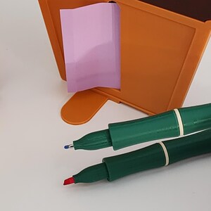 Peut inclure: Un organiseur de bureau orange avec un post-it rose et deux stylos verts avec des bandes blanches. Un stylo a une pointe bleue et l'autre une pointe rouge. L'organisateur est sur une surface blanche.