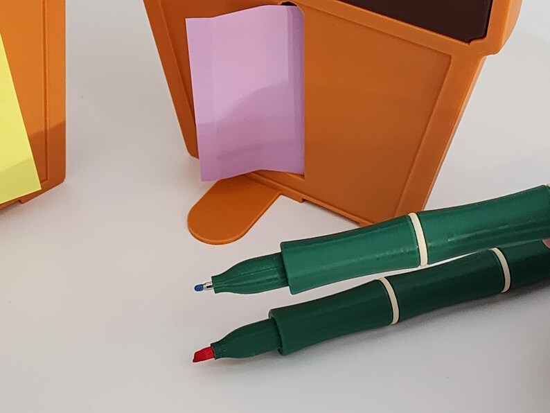 Peut inclure: Un organiseur de bureau orange avec un post-it rose et deux stylos verts avec des bandes blanches. Un stylo a une pointe bleue et l'autre une pointe rouge. L'organisateur est sur une surface blanche.