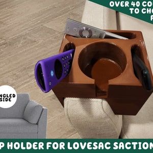 Puede incluir: Un portavasos de madera marrón diseñado para adaptarse a un sofá seccional lovesac. El portavasos tiene un portavasos, una ranura para un teléfono y una ranura para un mando a distancia. El portavasos está unido al lado del sofá.
