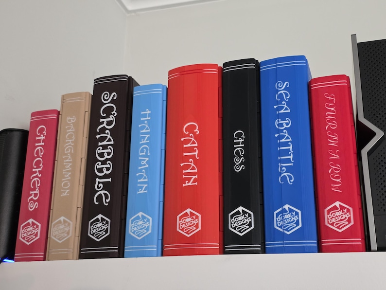 Peut inclure: Une collection de bo&icirc;tes de jeux de soci&eacute;t&eacute;, dont "Checkers", "Backgammon", "Scrabble", "Hangman", "Catan", "Chess", "Sea Battle" et "Four in a Row". Chaque bo&icirc;te est d'une couleur diff&eacute;rente et porte le titre du jeu sur le dos.