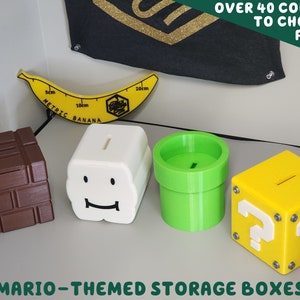 Op de afbeelding: Vier 3D-geprinte Mario-thema opbergdozen. De dozen zijn gevormd als een vraagtekenblok, een pijp, een baksteen en een personage uit het spel. De dozen zijn allemaal in verschillende kleuren, waaronder bruin, wit, groen en geel.