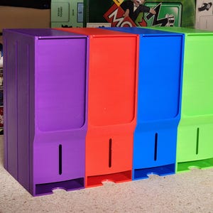 Puede incluir: Un conjunto de cuatro contenedores de almacenamiento coloridos en morado, rojo, azul y verde. Cada contenedor tiene forma rectangular con una ranura y una pequeña abertura en la parte inferior. Para organizar objetos.