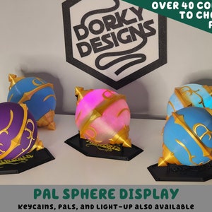 May include: Three 3D printed spheres with gold accents and different colored bases. The spheres are on black stands and have a design that resembles a magical orb. The text "PAL SPHERE DISPLAY" is at the bottom of the image. The text "keycains, PALS, AND LIGHT-UP ALSO AVAILABLE" is below the main text.