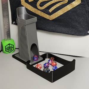 Puede incluir: Una torre de dados gris impresa en 3D con una bandeja negra, diseñada para juegos de mesa. La torre tiene un diseño arquitectónico alto con ranuras verticales. Varios dados de múltiples caras están en la bandeja. Una regla y un cubo verde con el texto "Dorky Designs" están en el fondo.