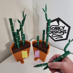 Peut inclure: Deux pots de plantes en bambou orange et marron avec des tiges et des feuilles de bambou vertes. Les pots ont des notes adh&eacute;sives jaunes et roses attach&eacute;es. Un stylo vert avec un motif de bambou est tenu dans une main. Le logo "Dorky Designs" est en arri&egrave;re-plan.