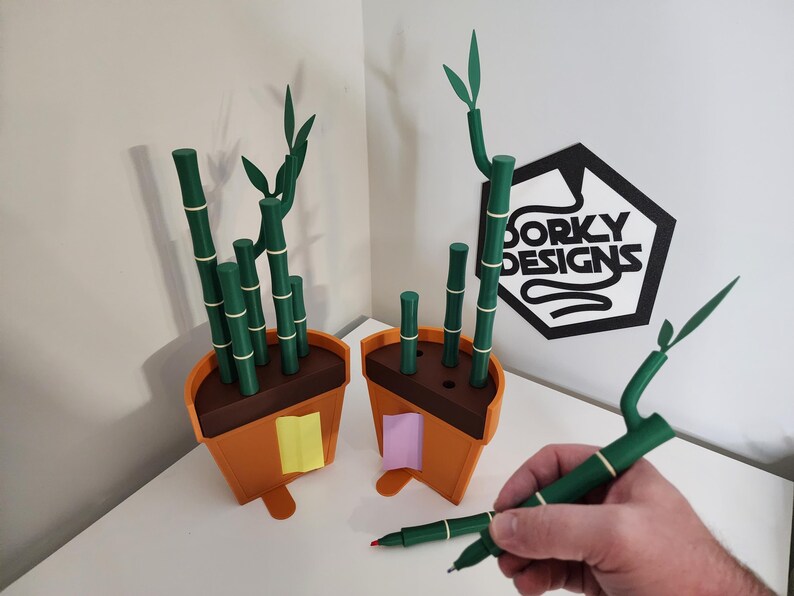 Peut inclure: Deux pots de plantes en bambou orange et marron avec des tiges et des feuilles de bambou vertes. Les pots ont des notes adh&eacute;sives jaunes et roses attach&eacute;es. Un stylo vert avec un motif de bambou est tenu dans une main. Le logo "Dorky Designs" est en arri&egrave;re-plan.