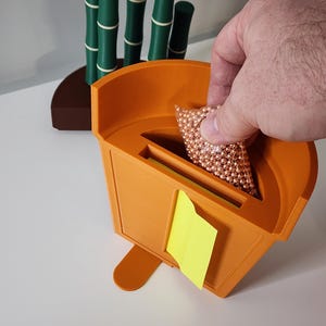 Peut inclure: Un conteneur orange imprim&eacute; en 3D avec une fente et un post-it jaune. Une main place un sac de petites sph&egrave;res de couleur cuivre dans la fente. D&eacute;cor en forme de bambou en arri&egrave;re-plan.