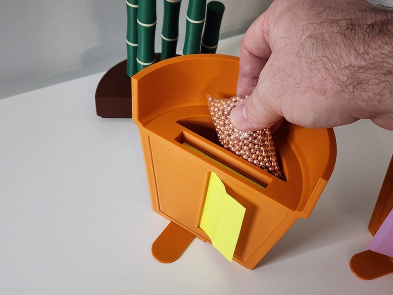 Peut inclure: Un conteneur orange imprim&eacute; en 3D avec une fente et un post-it jaune. Une main place un sac de petites sph&egrave;res de couleur cuivre dans la fente. D&eacute;cor en forme de bambou en arri&egrave;re-plan.