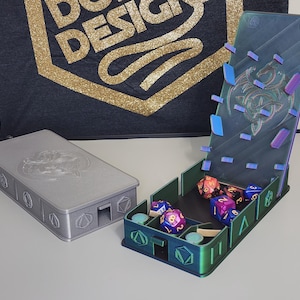Könnte beinhalten: Ein 3D-gedruckter Würfelturm mit einem klaren, schillernden Deckel und einem Drachenmotiv. Der Turm hat Fächer für Würfel und einen Schlitz zum Rollen. Der Turm steht auf einem Tisch mit einer grauen 3D-gedruckten Box.