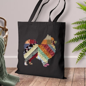 May include: Black tote bag featuring a colorful patchwork dog design. The dog silhouette is composed of various fabric patterns in shades of red, orange, blue, and green. The bag has black handles and is displayed against a neutral background.