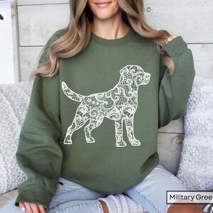 May include: Military green sweatshirt featuring a white lace-patterned dog silhouette. The dog design is detailed and covers the entire body of the dog. The sweatshirt is a solid color and has a crew neck.
