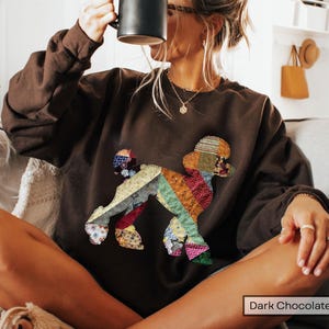 May include: Dark chocolate-colored sweatshirt featuring a colorful patchwork poodle design. The poodle is made of various fabric scraps in shades of red, blue, green, and yellow. The sweatshirt has a crew neck and long sleeves.