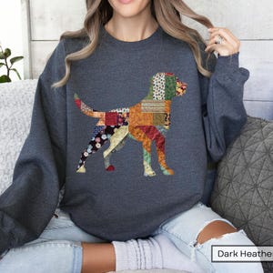 May include: Dark heather gray sweatshirt featuring a colorful patchwork dog design. The dog silhouette is composed of various fabric patterns in shades of red, orange, blue, and green. The sweatshirt has long sleeves and a crew neck.