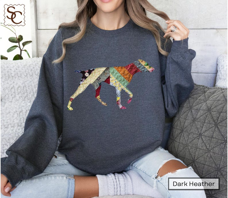 May include: Dark heather grey sweatshirt with a patchwork dog design. The dog is made of various fabric patterns in red, yellow, green, and blue. The sweatshirt has a crew neck and long sleeves.