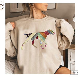May include: Sand-colored sweatshirt featuring a colorful patchwork dog design. The dog is depicted in a walking pose, with the patchwork design incorporating various colors and patterns. The sweatshirt has long sleeves and a relaxed fit, suitable for casual wear.